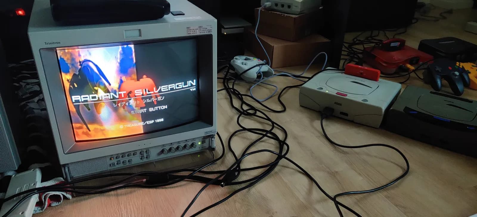 A white/cream Sega Saturn Model 2 (with a red cartridge in its slot) running the Radiant Silvergun title screen (Treasure/ESP, 1998) on a Sony PVM-14L2 broadcast monitor, a white Saturn controller and other consoles on the wood floor during the post-recap smoke test.