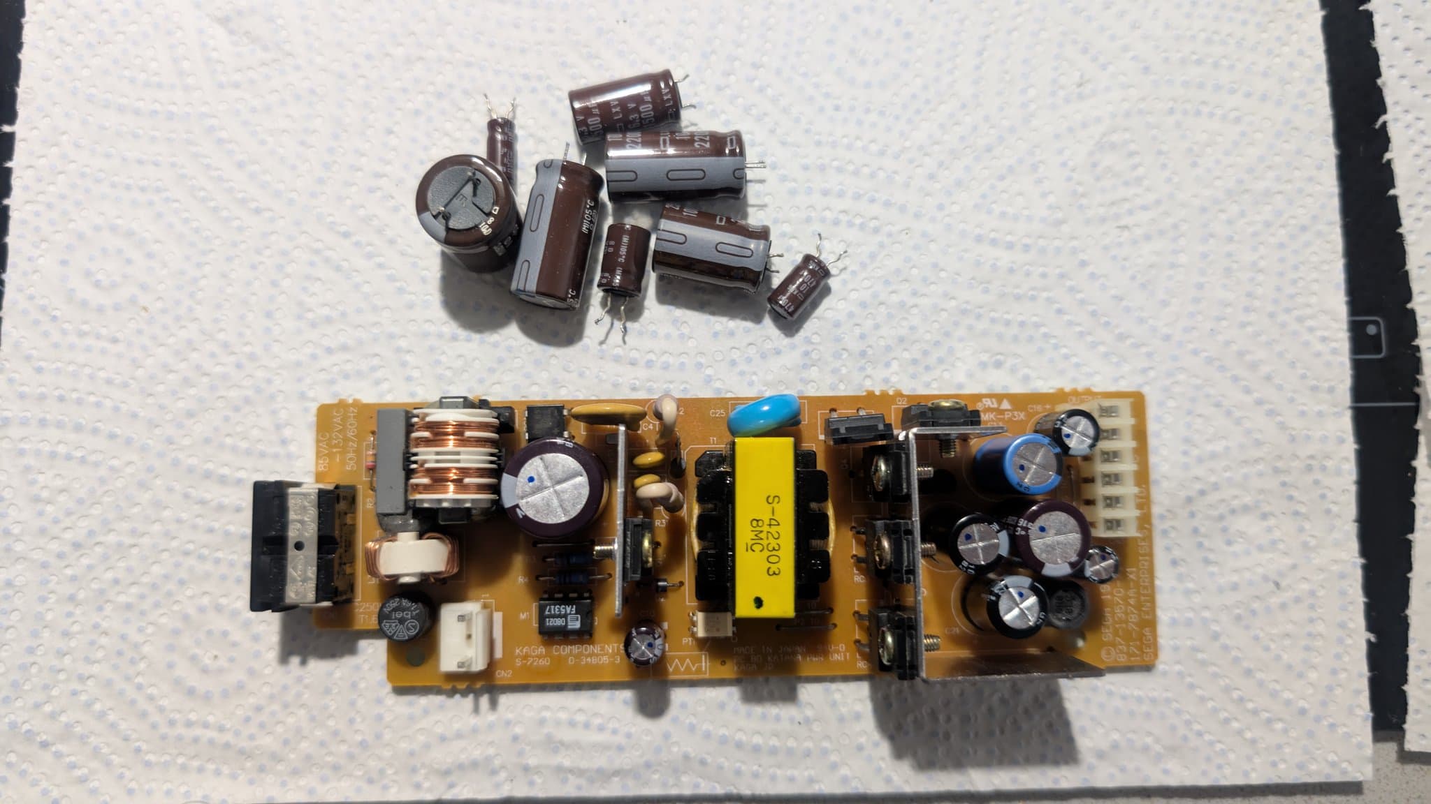 Dreamcast VA0 PSU board with recapped electrolytic capacitors; loose old capacitors displayed above the repaired power supply unit.