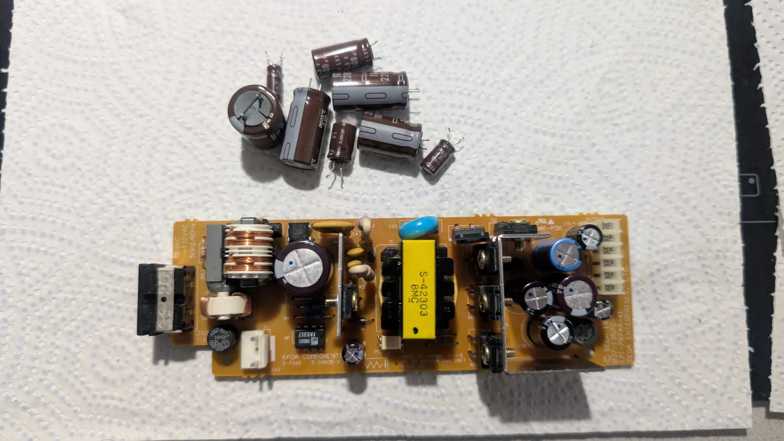 Dreamcast VA0 PSU board with recapped electrolytic capacitors; loose old capacitors displayed above the repaired power supply unit.