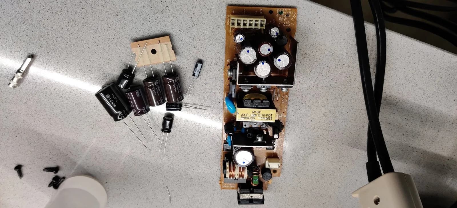 The Sega Dreamcast VA0 PSU board (marked PSM168) before recapping, with its original capacitors still fitted, beside the new replacement capacitors, a TO-220 regulator on a small pad, a diode and case screws on a speckled stone countertop.