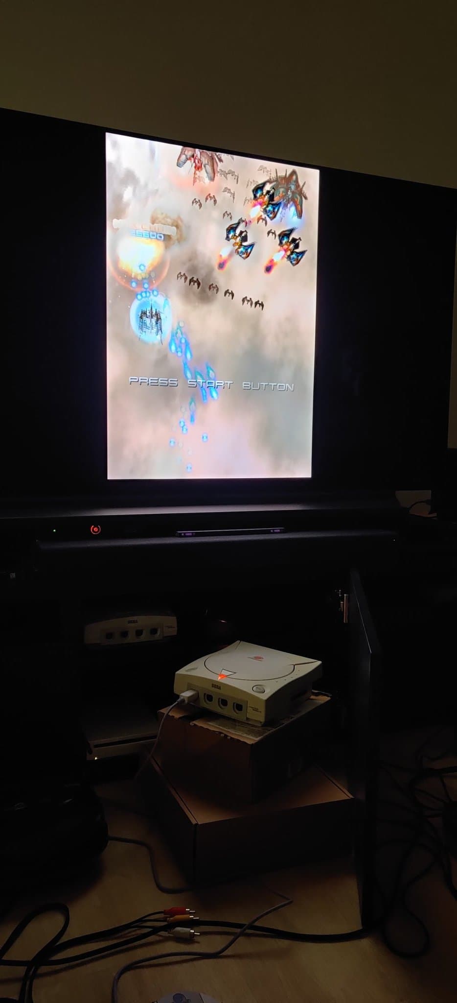 Sega Dreamcast VA0 displaying Ikaruga title screen via GDEMU mod, connected to display; console visible on shelf below.