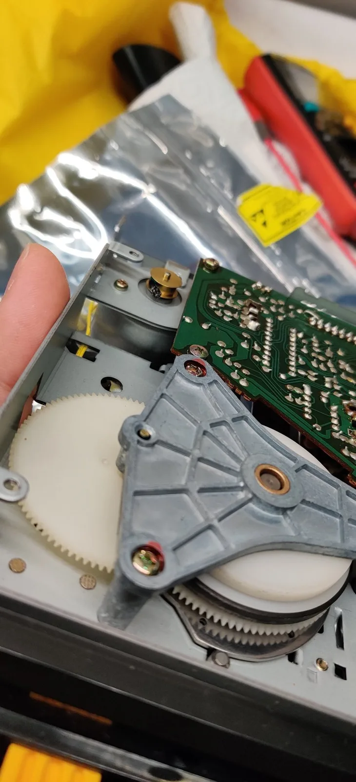 Close-up of the Famicom Disk System drive mechanism before refurbishment, held in hand: the small control board is bolted to the metal casing beside the motor that drives the belt, and the remnants of the old perished belt are melted onto the brass motor pulley, before a new belt was fitted and the old residue cleaned off; large white plastic gears sit alongside.