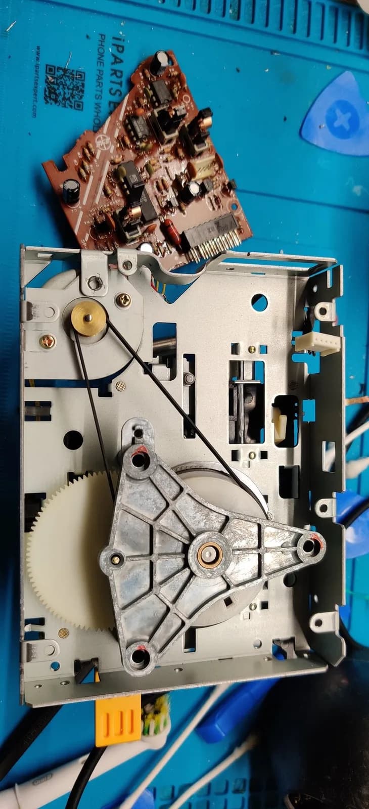 Famicom Disk System drive mechanism on a blue work mat, the newly fitted black rubber drive belt looping from the brass motor pulley down to the large flywheel, beside a cream toothed gear and a grey die-cast bracket on the bare silver metal chassis; the maroon control board detached above.