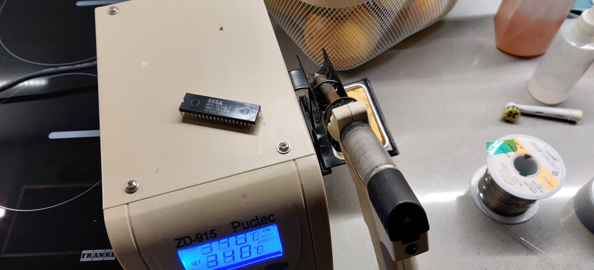 The original Sega Mega-CD 2 BIOS chip, freshly desoldered, resting on top of a ZD-915 Puctec desoldering station, with a soldering iron and solder spool nearby during the repair.