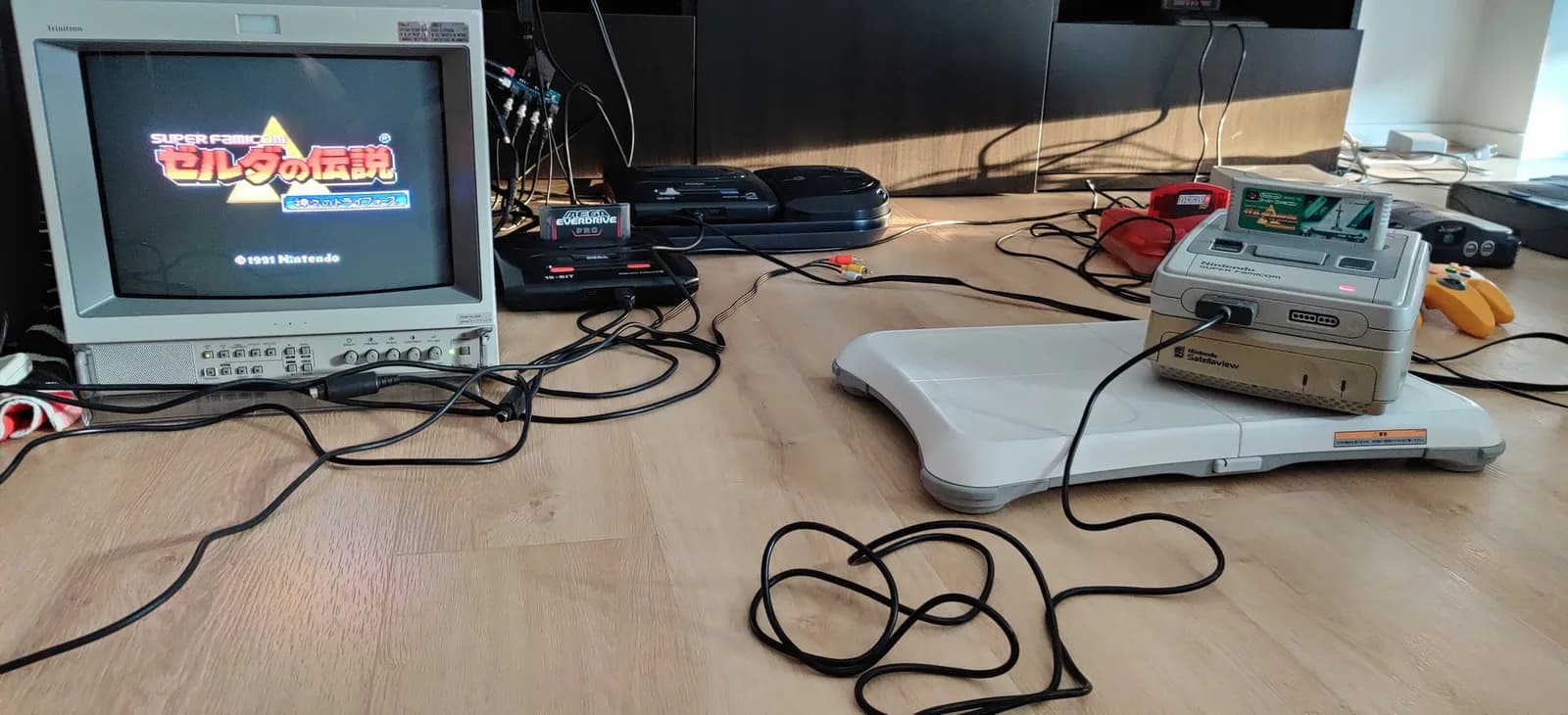 Repaired Nintendo Super Famicom sitting on top of a Nintendo Satellaview base, running The Legend of Zelda: A Link to the Past on a Sony PVM-14L2; a Sega Mega Drive with a Mega Everdrive Pro cartridge, other consoles, controllers and cables around it on a wood surface.