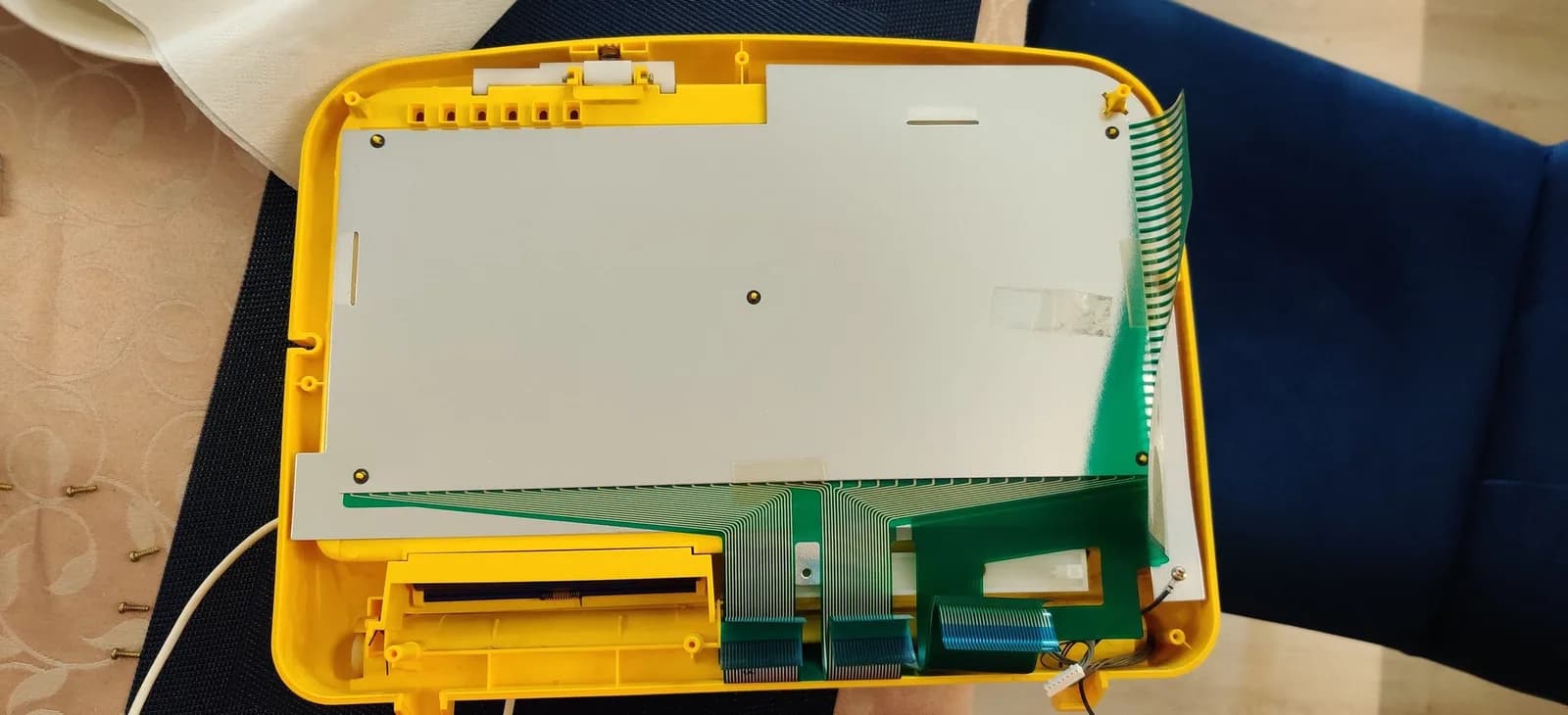 Inside of the yellow top half of a Sega Pico shell laid face-down, showing the shielding, green flexible ribbon cables fanning out and folding over the edge, and a small wire connector.