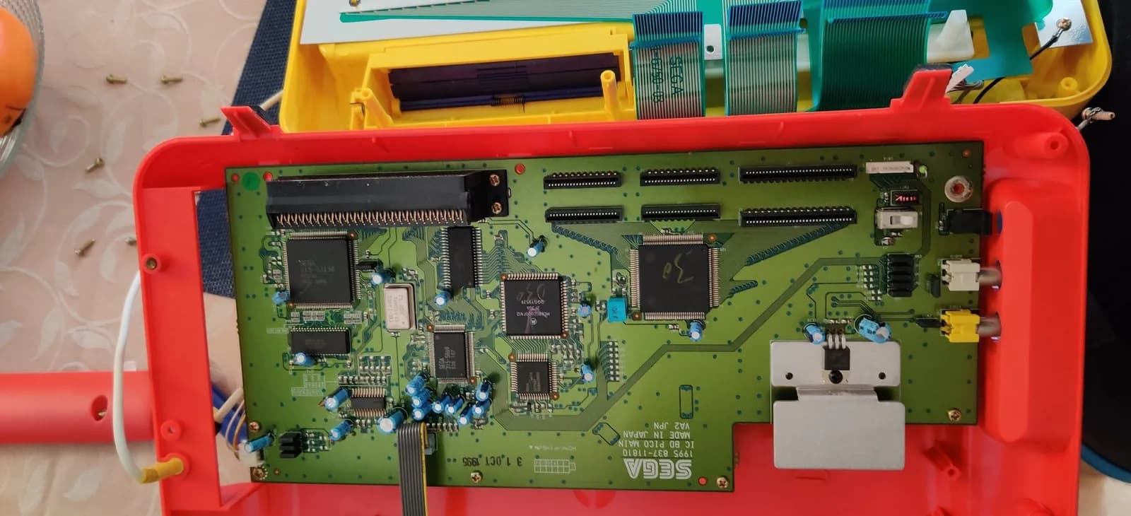 First-revision Sega Pico mainboard seated in the red lower shell before the recap, with its original electrolytic capacitors, ICs, RCA AV jacks and the regulator screwed into a metal heatsink.
