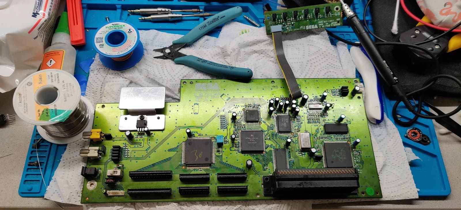 Sega Pico mainboard with its recap completed, on a blue silicone mat surrounded by a solder spool, a copper desoldering braid on a blue spool, a bottle of super glue, flush cutters and a soldering iron handpiece. The green PCB shows its ICs and the new capacitors.
