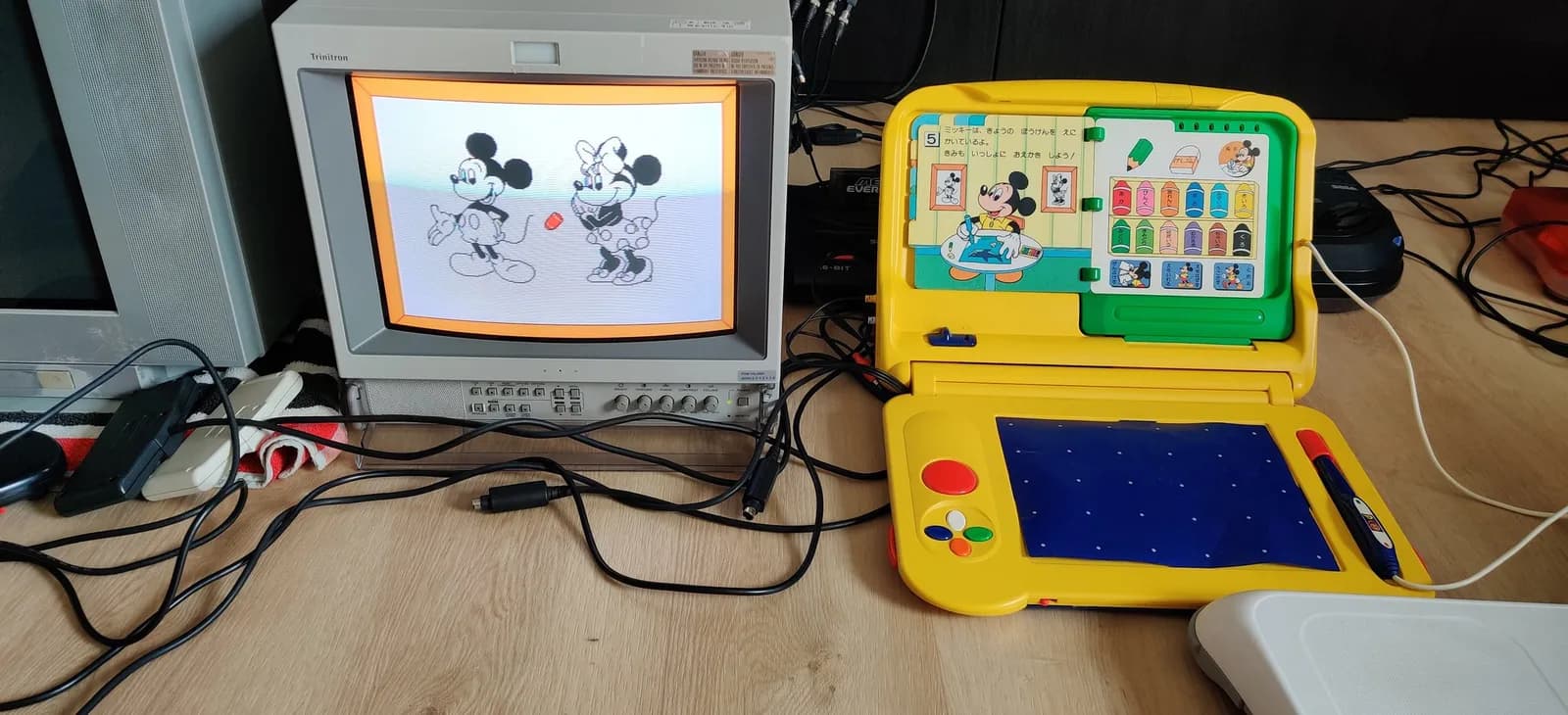 Recapped yellow Sega Pico, a clamshell tabletop console, open on the floor beside a Sony PVM-14L2 CRT showing a Mickey Mouse game. A Storyware picture book is slotted inside the clamshell's top-half with its lower edge in the cartridge slot, and the console's lower-half has a blue drawing tablet, coloured buttons and a dark blue tethered Magic Pen seated in its orange holder on the shell.