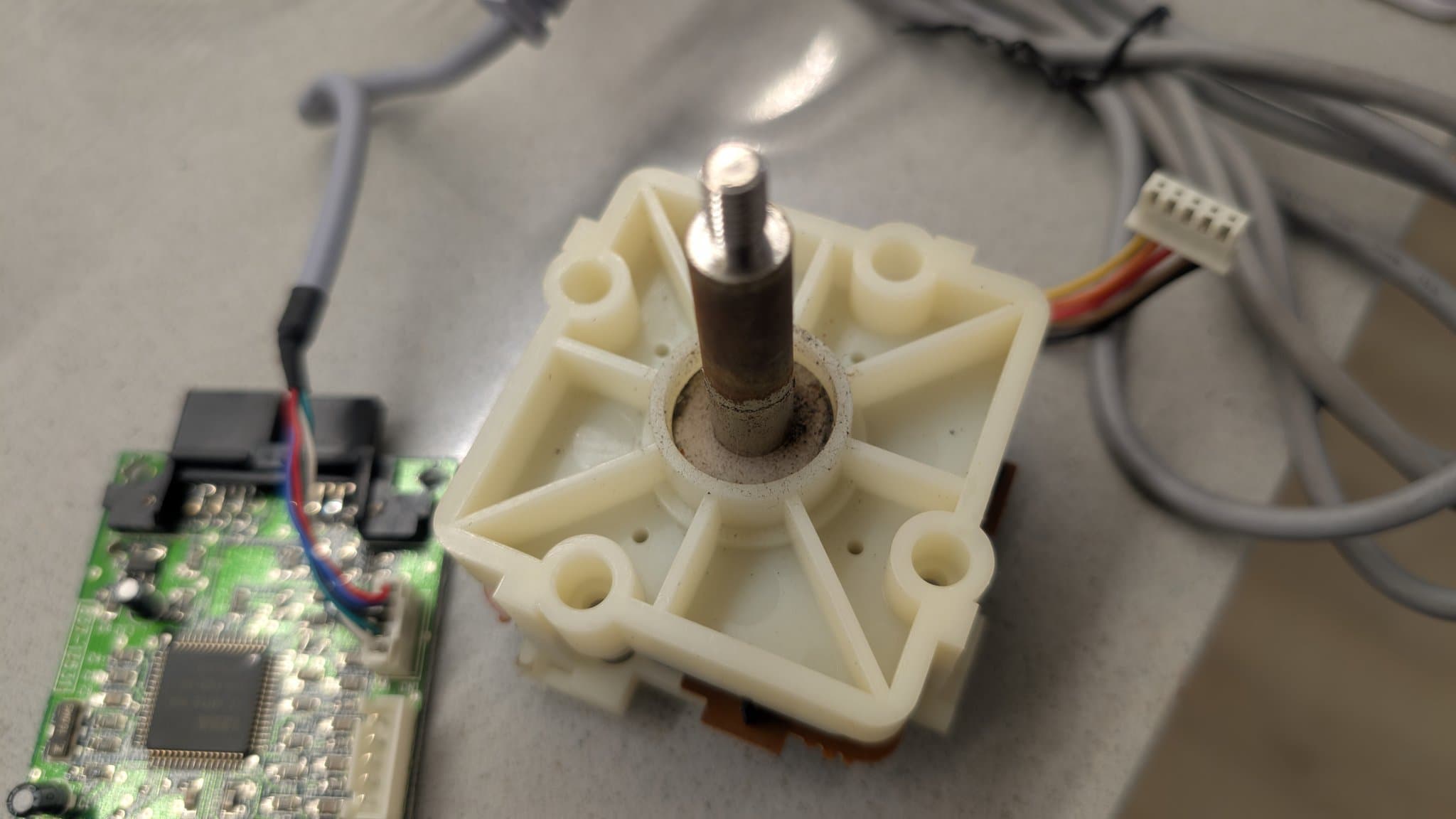 The Dreamcast arcade stick's joystick mechanism with its dusty metal pivot shaft and cream plastic housing, resting on the bench beside a separate green control PCB and the plug-in connector that links the stick's wiring to the console's controller board.