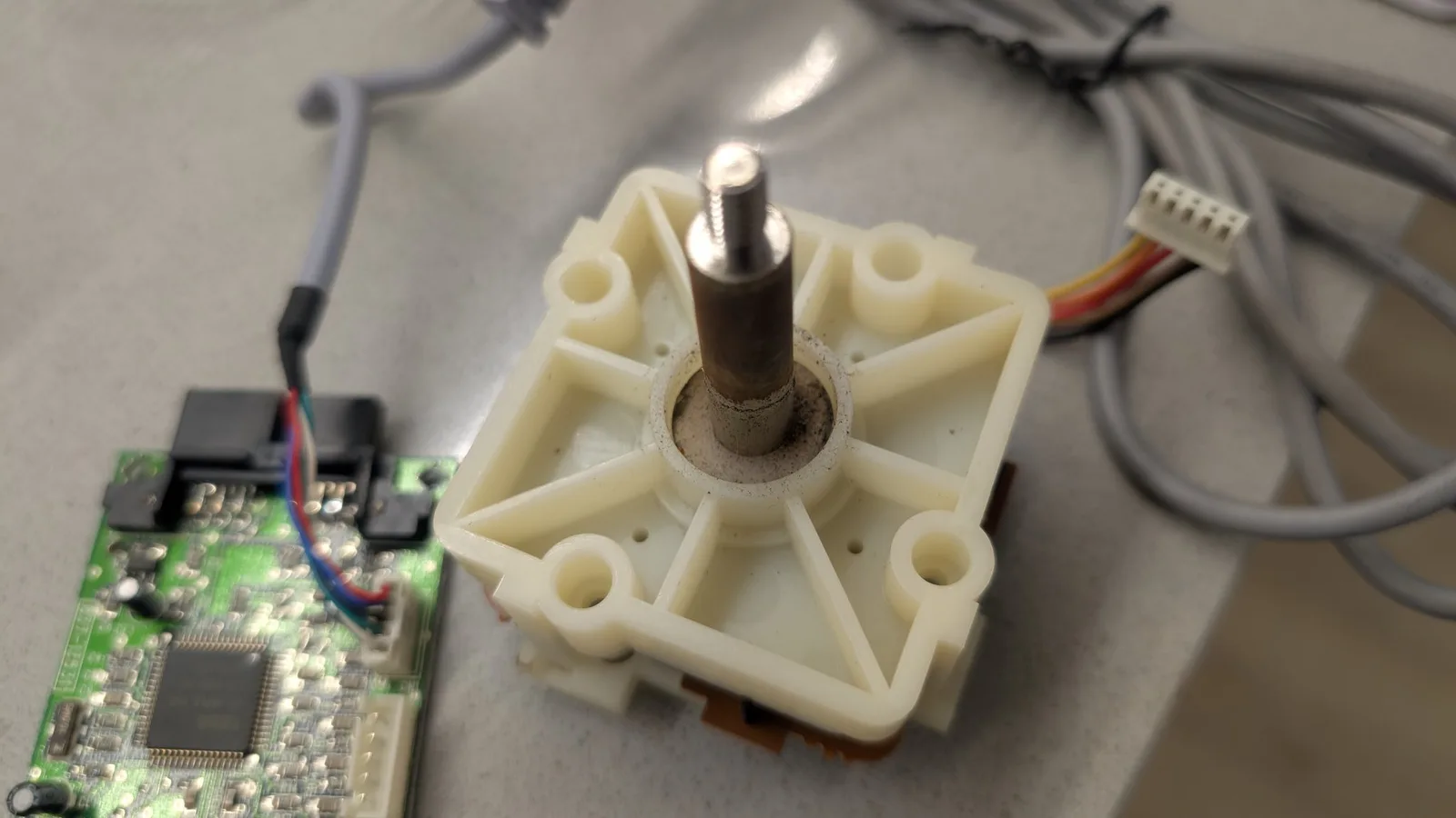 The Dreamcast arcade stick's joystick mechanism with its dusty metal pivot shaft and cream plastic housing, resting on the bench beside a separate green control PCB and the plug-in connector that links the stick's wiring to the console's controller board.