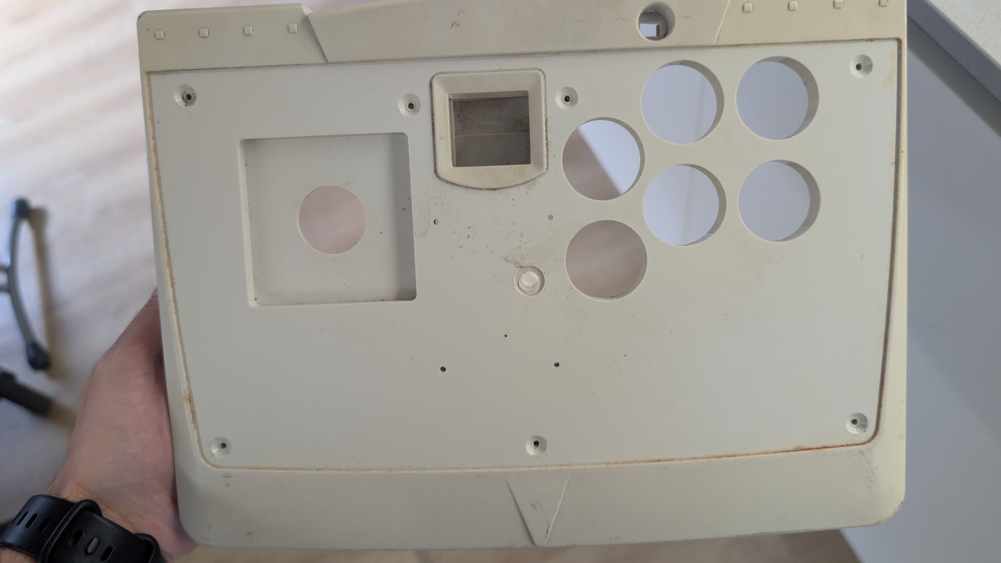 The yellowed top housing of the Dreamcast arcade stick held in hand, with the square joystick opening, six round button holes and the rectangular control-panel cutout, rust staining at the edges.