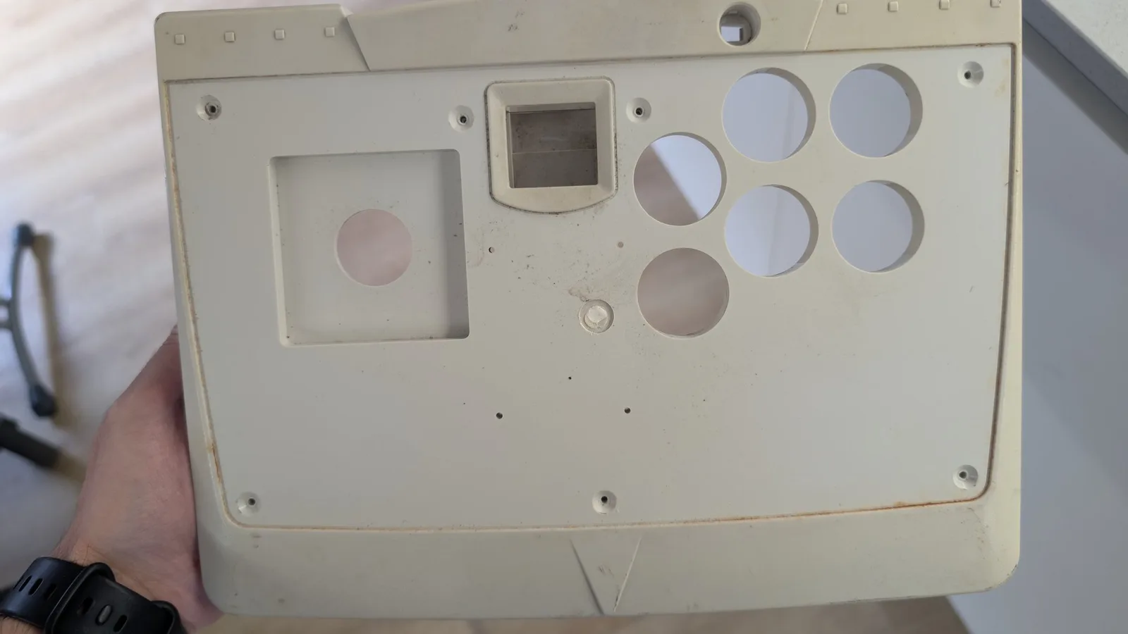 The yellowed top housing of the Dreamcast arcade stick held in hand, with the square joystick opening, six round button holes and the rectangular control-panel cutout, rust staining at the edges.