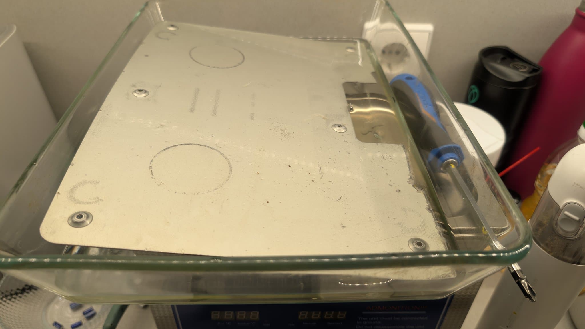 The Dreamcast arcade stick's painted metal base plate and a steel shield plate soaking in Evaporust rust-removal solution in a clear glass container, a screwdriver resting against the outside of it.