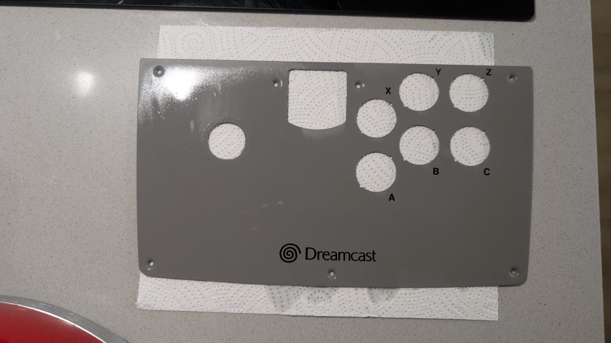 The Dreamcast arcade stick's bare metal control faceplate after cleaning, the Dreamcast logo and the empty button holes labeled X, Y, Z, A, B, C around the joystick opening, on paper towel.