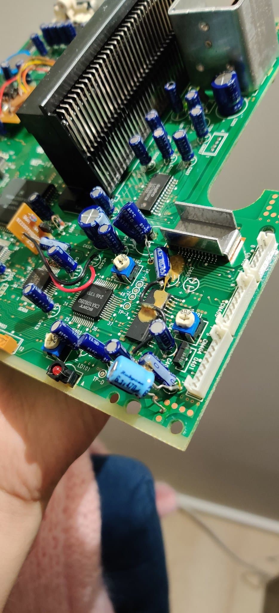 Mega-CD 2 PCB held in hand showing the heatsink, blue capacitors and ICs, with the various factory bodges on the side of the board behind the connector that slots into the Mega Drive.