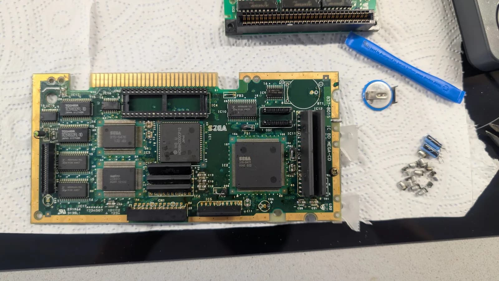Sega Mega-CD 1 PCB during recap, showing multiple leaking capacitors removed, with soldering iron and desoldered components on white work mat.