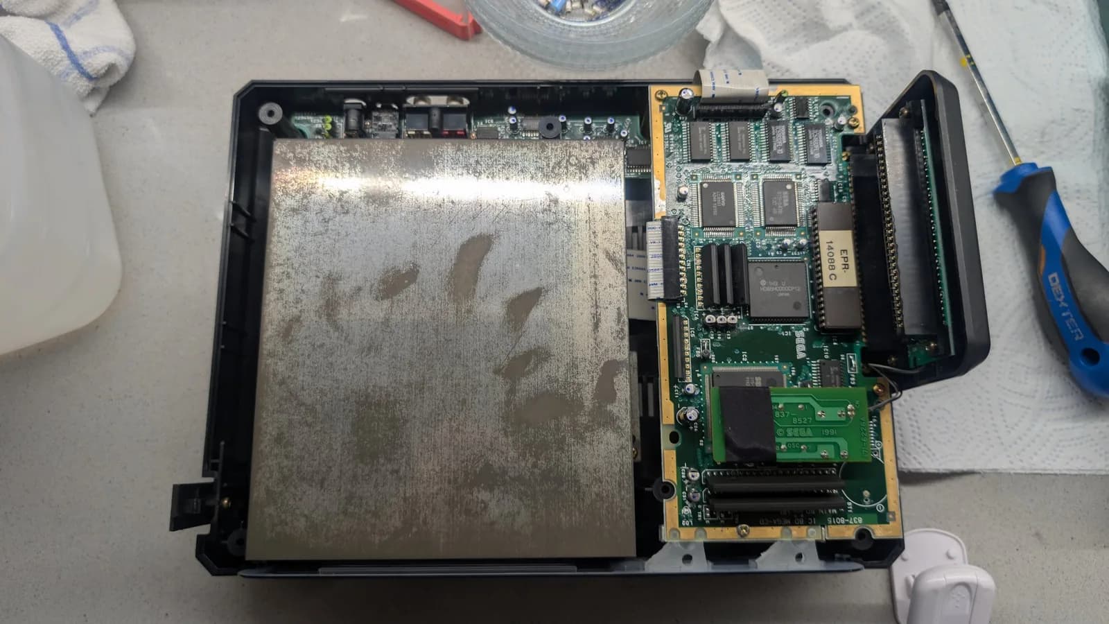 Sega Mega-CD 1 interior disassembled showing large metallic shielding, circuit boards with IC chips, and yellow/green PCBs during restoration diagnostics.