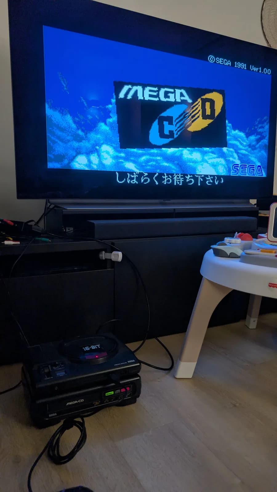 Sega Mega-CD 1 displaying BIOS startup screen with "MEGA" logo and Japanese wait message; console and controller visible below TV.