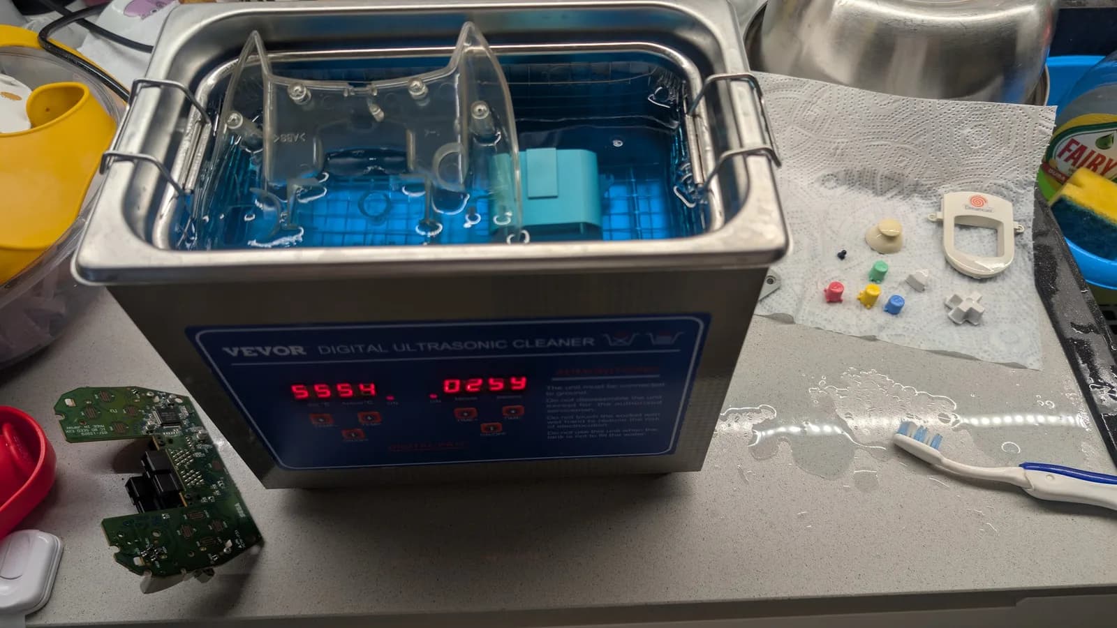Dreamcast controller components soaking in VEVOR ultrasonic cleaner displaying 55.54°C and 0259 settings, with PCB and disassembled parts on work surface.