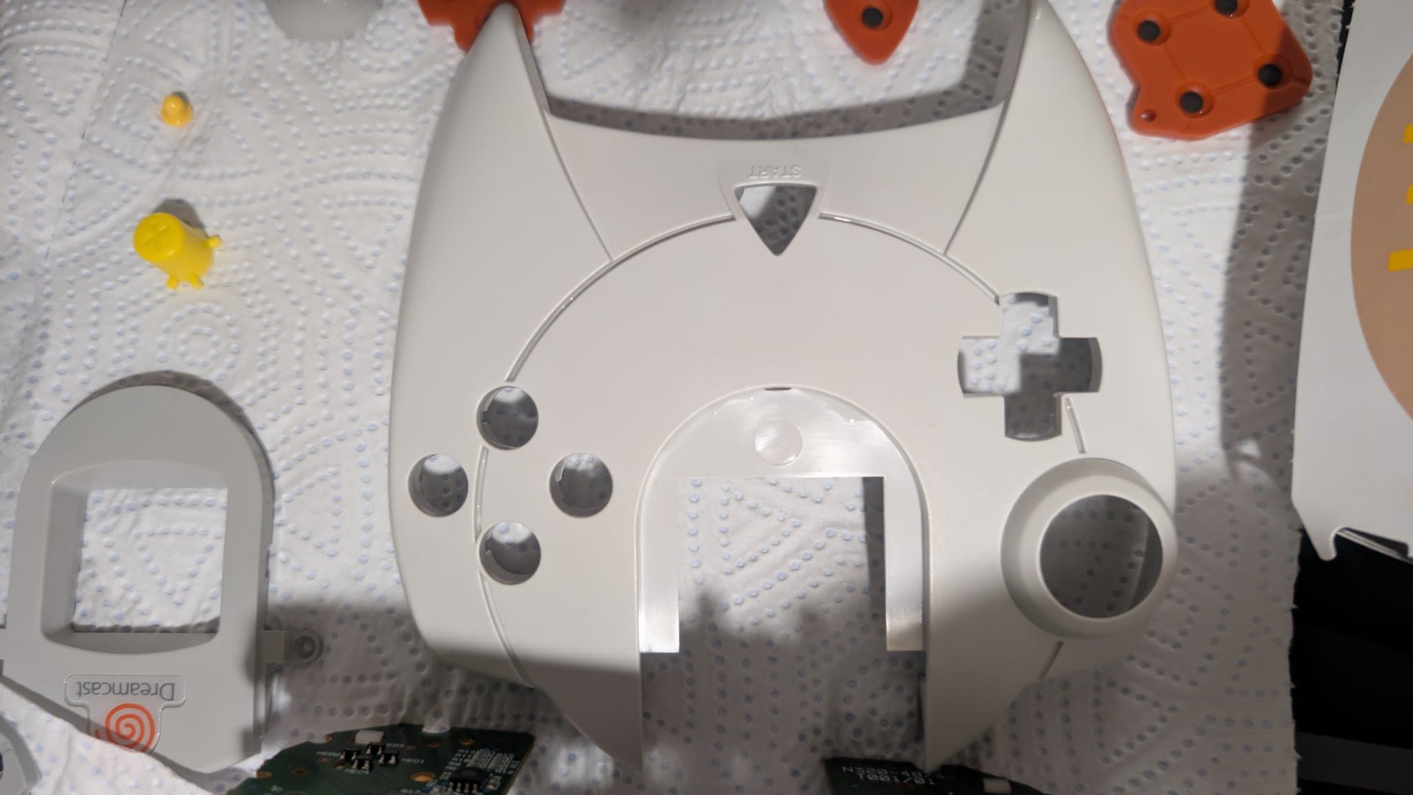 Cleaned Dreamcast controller front shell on a white surface, the bare cutouts for the d-pad, face buttons and analog stick and the VMU window visible, with the orange rubber button pads and loose buttons resting separately nearby.