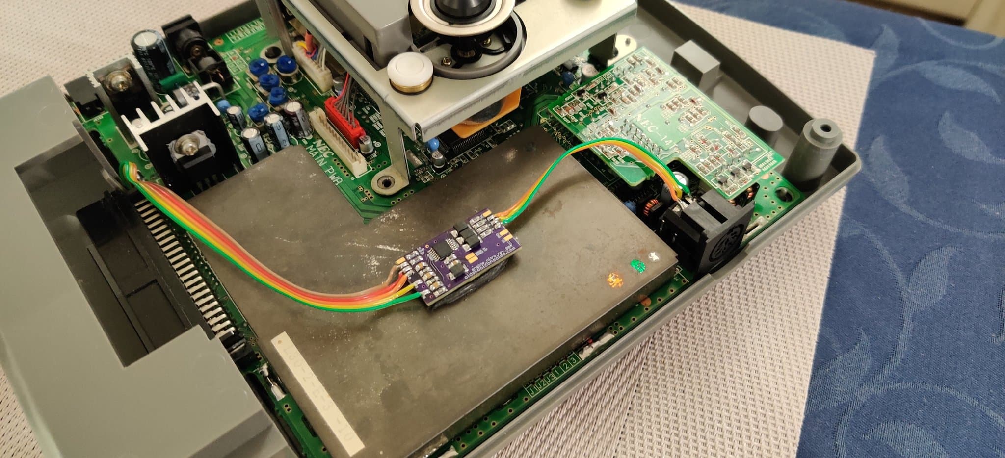 Interior of the NEC PC Engine Super CD-ROM2 showing the recapped mainboard (the power circuitry is part of it, there is no separate PSU board), an RGB amplifier module with a rainbow ribbon cable, and an 8-pin DIN connector being installed during the refurbishment.