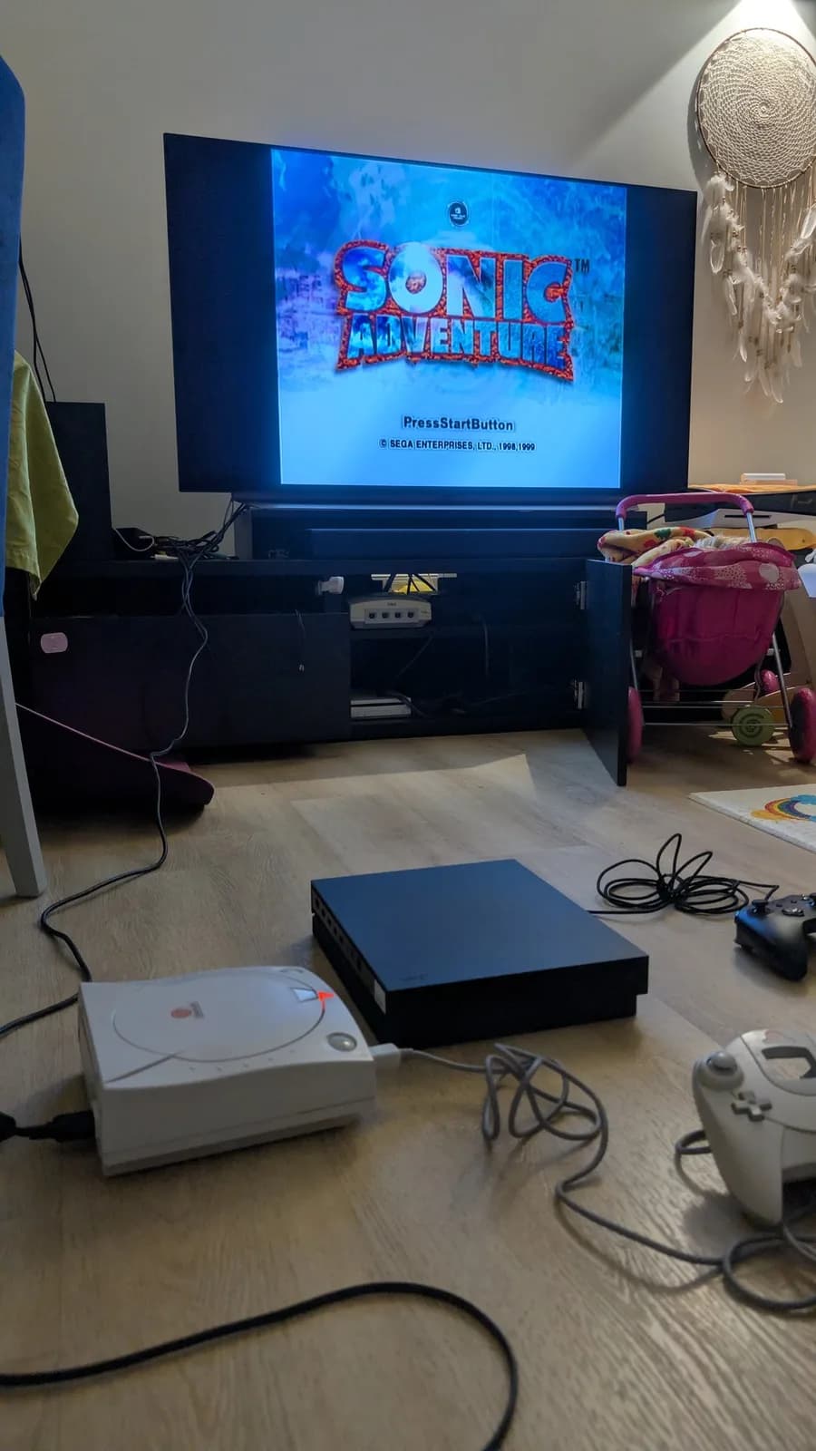 Sega Dreamcast VA0 startup screen showing Sonic Adventure burn-in test, with white Dreamcast and black console visible on wooden floor.