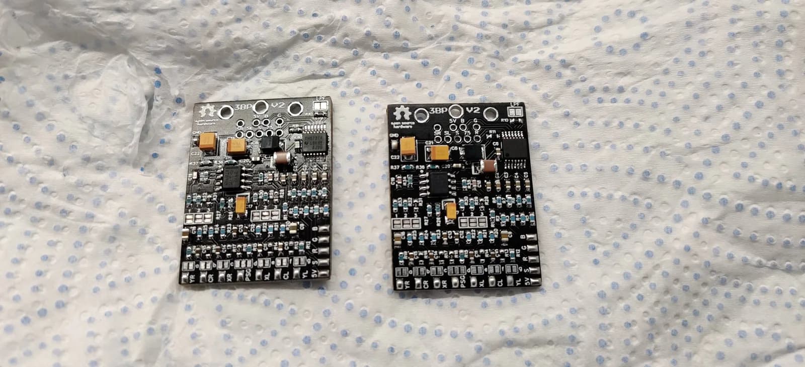 The two Triple Bypass (3BP) V2 boards fully assembled, side by side on a paper towel, the orange tantalum capacitors, resistors and ICs all fitted with clean solder joints.