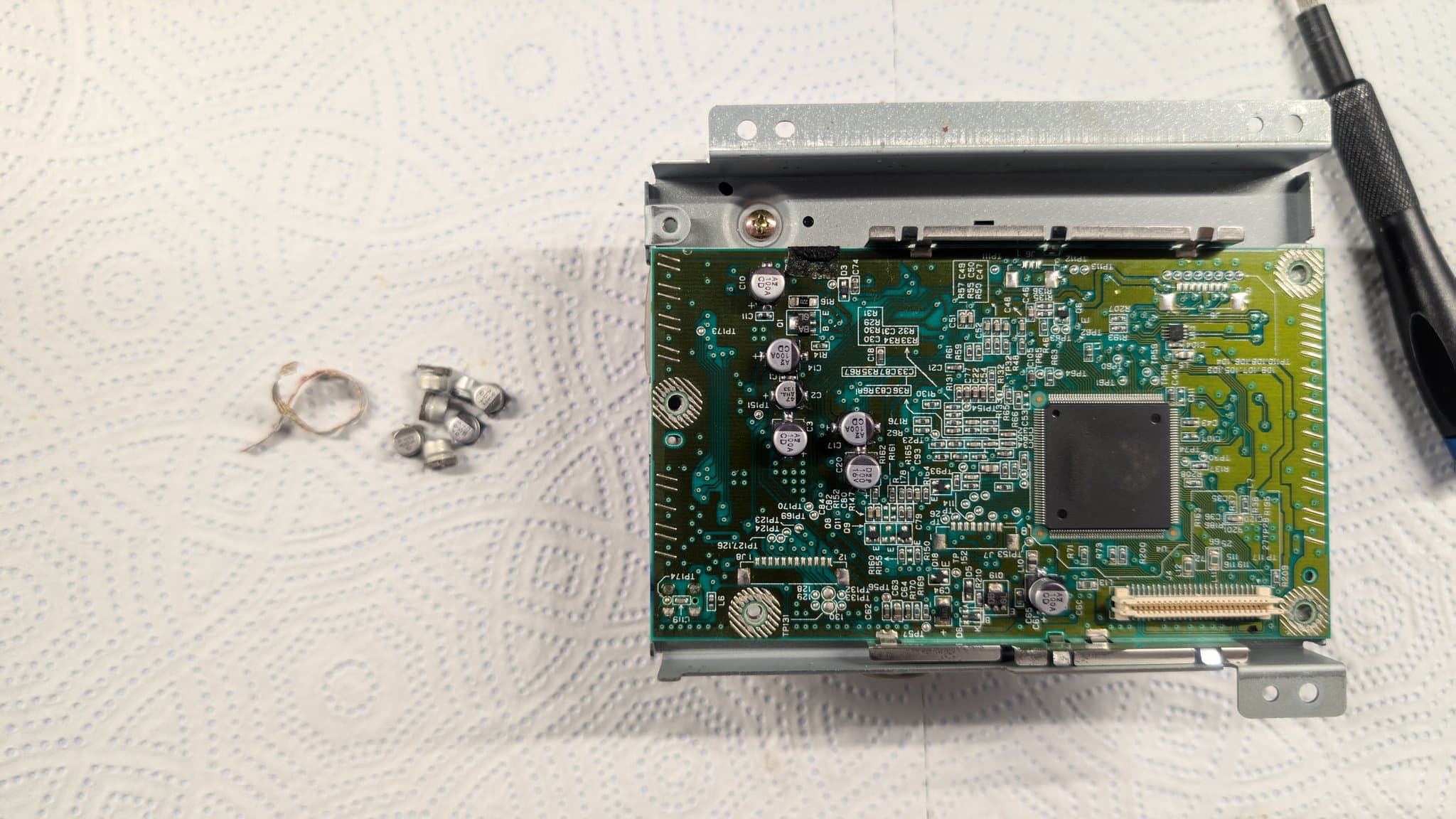 The Dreamcast GD-ROM drive board on its metal bracket, the old electrolytic capacitors removed and laid out on the mat beside it during the recap.