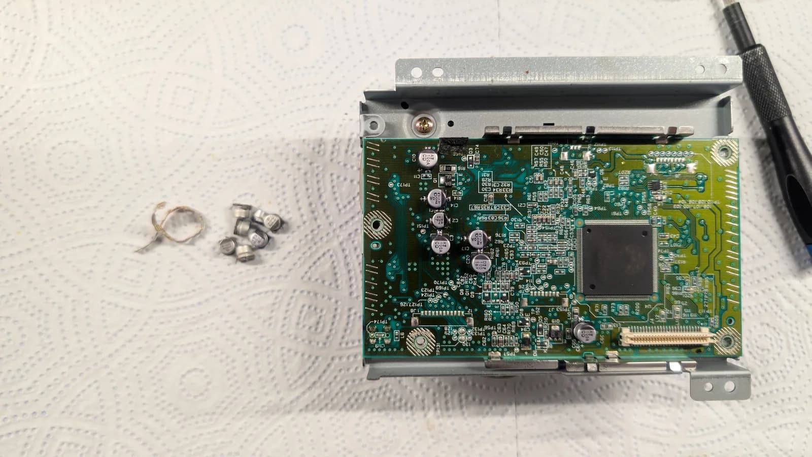 The Dreamcast GD-ROM drive board on its metal bracket, the old electrolytic capacitors removed and laid out on the mat beside it during the recap.