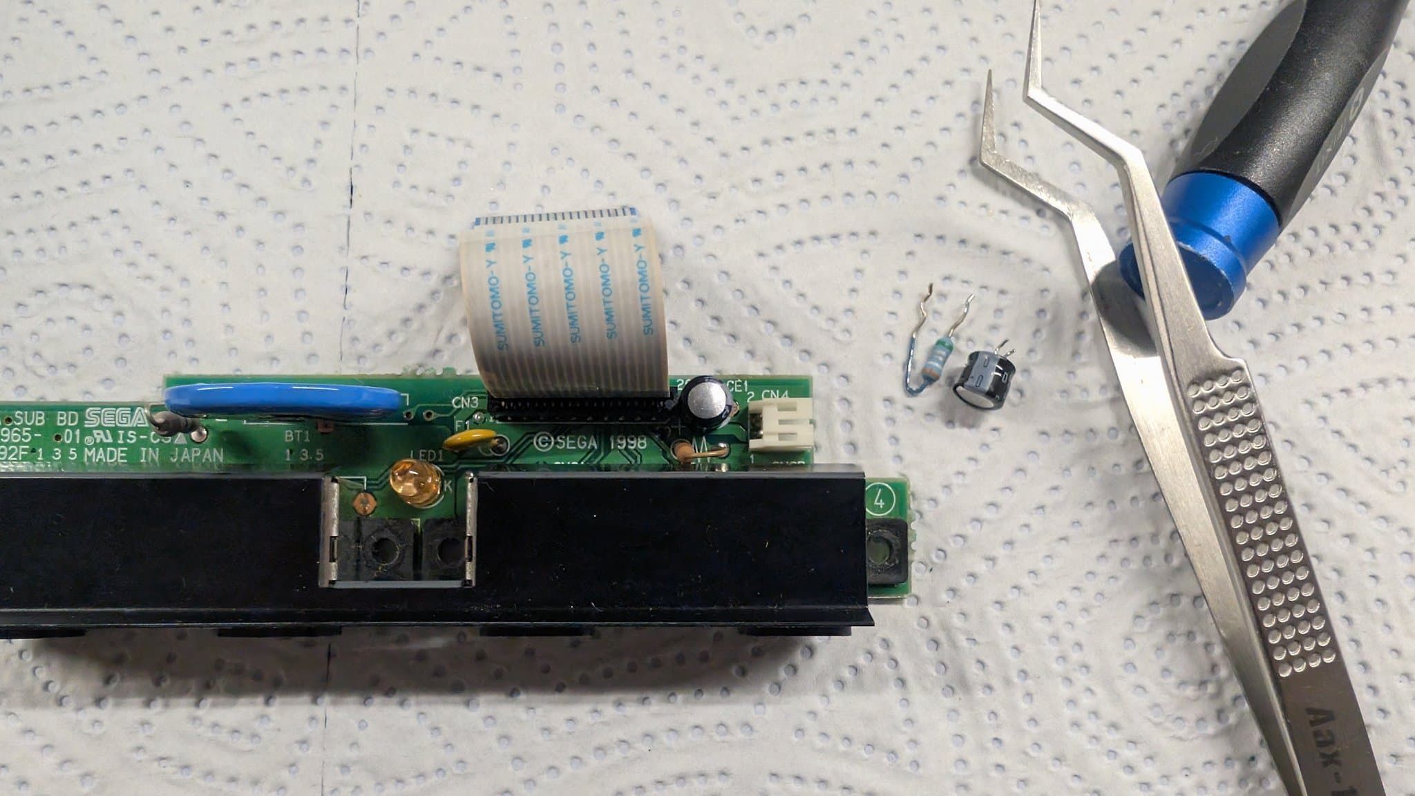 The Dreamcast controller-port sub-board with its old capacitor and fuse removed onto the mat beside the tweezers, replaced with a new capacitor and a resettable fuse.