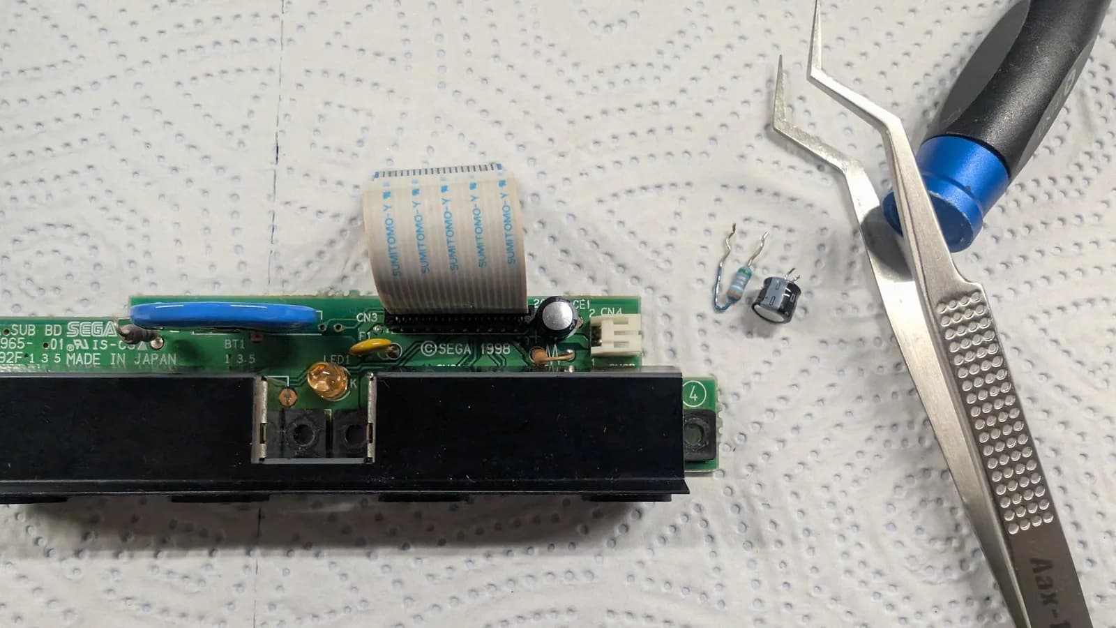 The Dreamcast controller-port sub-board with its old capacitor and fuse removed onto the mat beside the tweezers, replaced with a new capacitor and a resettable fuse.