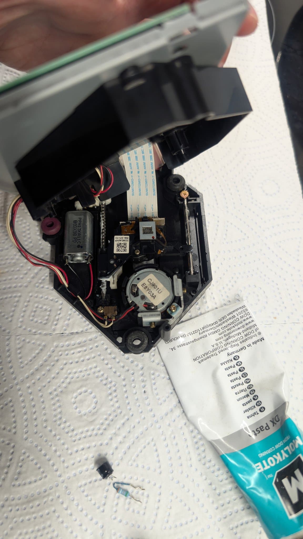 The Dreamcast GD-ROM optical-drive mechanism opened for lubrication, the spindle motor and ribbon cable visible, with a tube of Molykote paste on the mat.