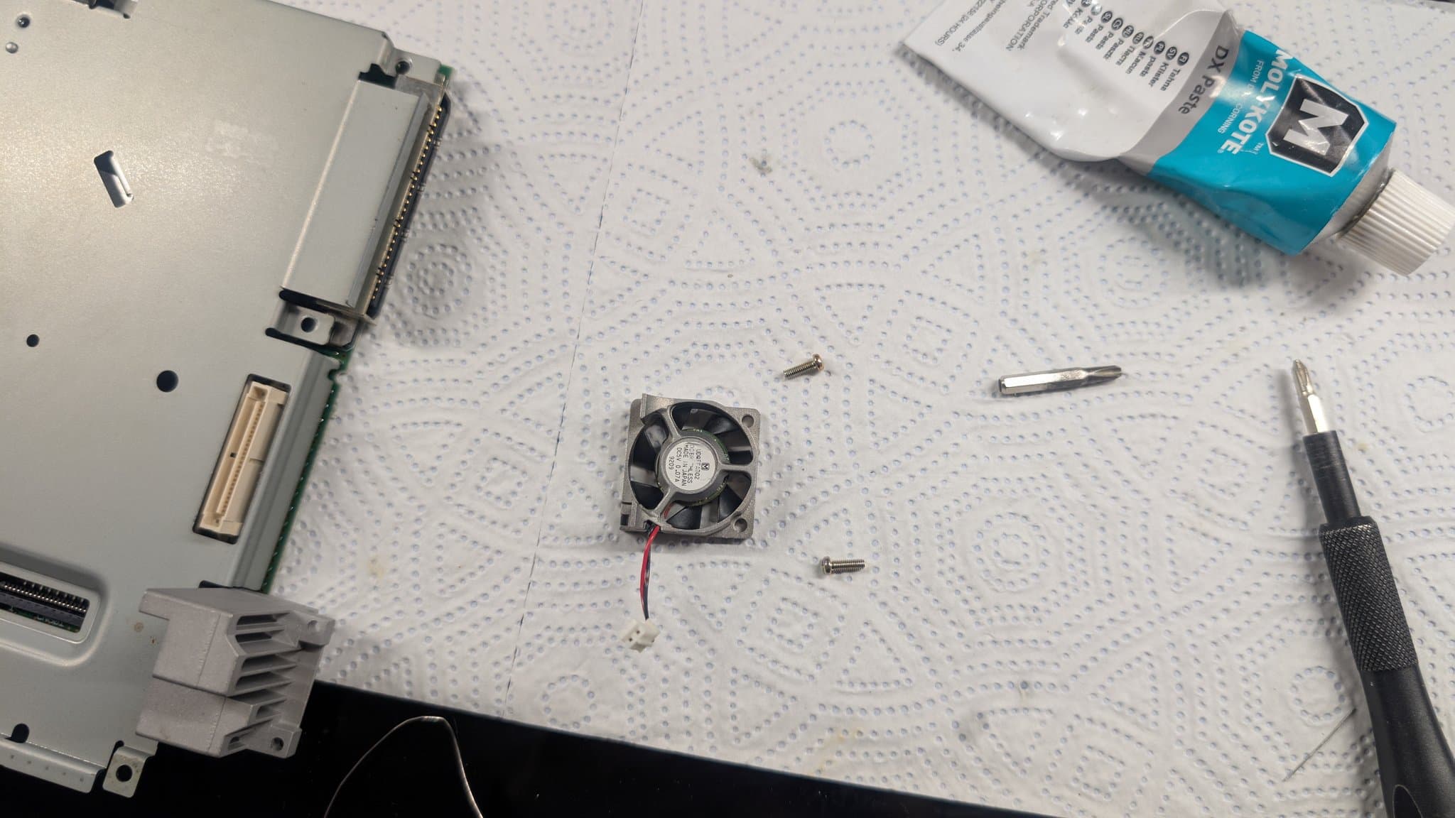 Dreamcast VA0 teardown with the motherboard and its metal shielding, the cooling fan with red and black wires, screws, a screwdriver and a tube of Molykote paste on the mat.