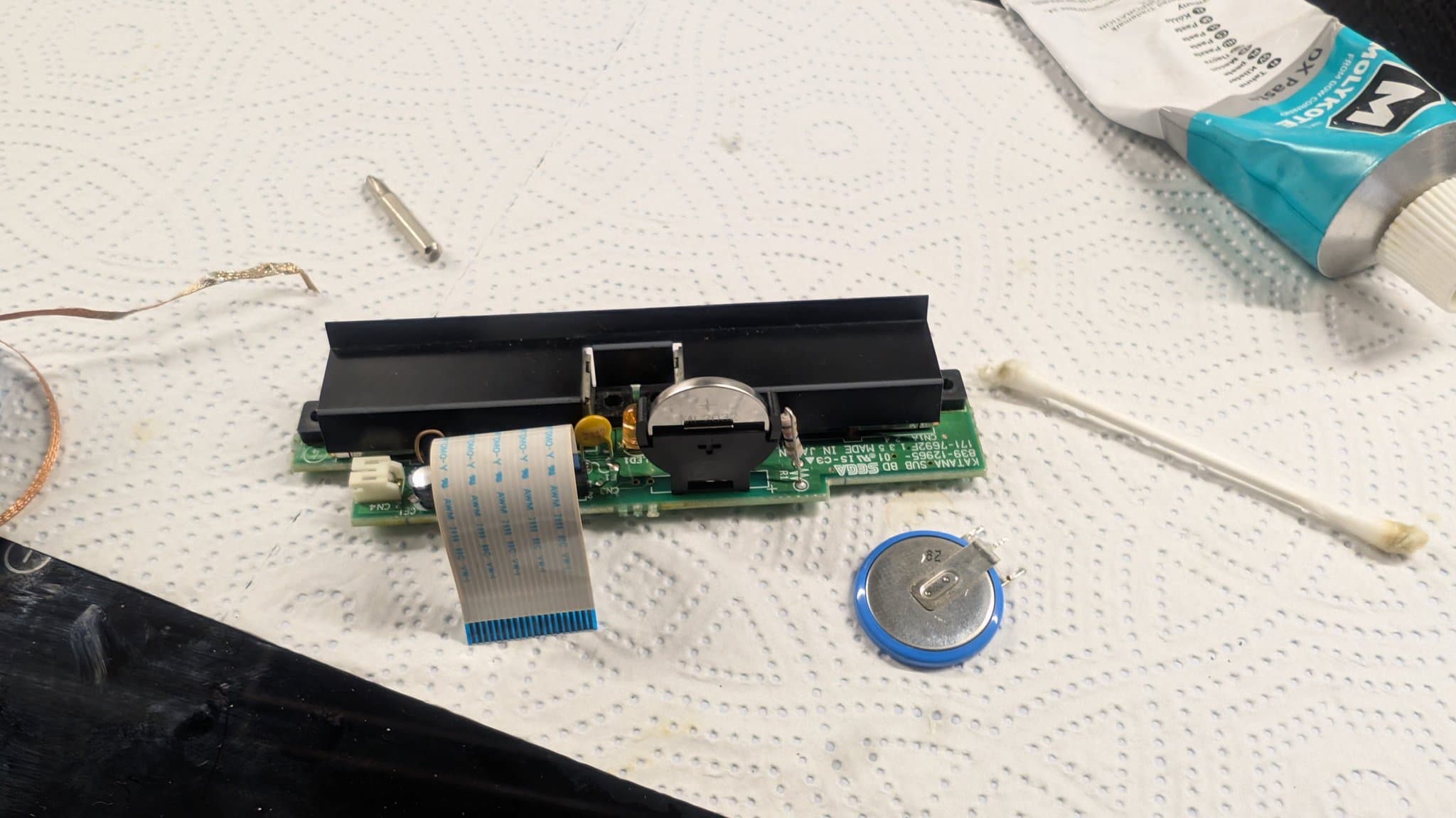 The Dreamcast controller-port sub-board (KATANA-SUB) with its ML2032 clock battery in the holder and the ribbon cable attached, the removed old cell and a cotton swab beside it.