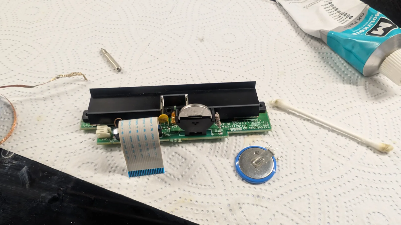 The Dreamcast controller-port sub-board (KATANA-SUB) with its ML2032 clock battery in the holder and the ribbon cable attached, the removed old cell and a cotton swab beside it.