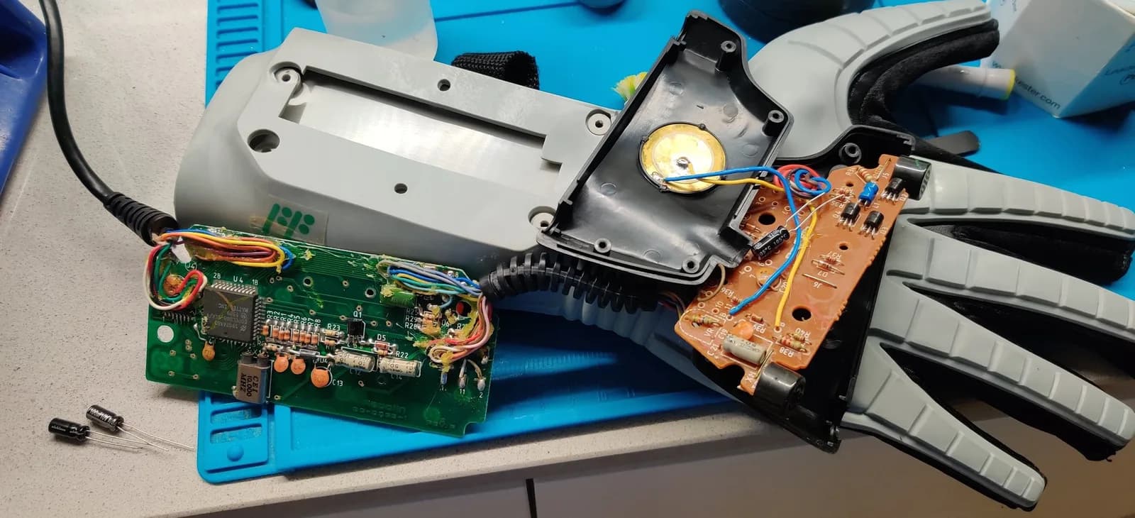 Disassembled Nintendo Power Glove on a blue work mat: the green main PCB with its square IC at left, the copper/orange daughterboard at right with a new capacitor resting on top ready to replace the old one, two new black capacitors on the mat waiting to go into the green board, plus the grey glove housing and a black housing with a round gold disc.