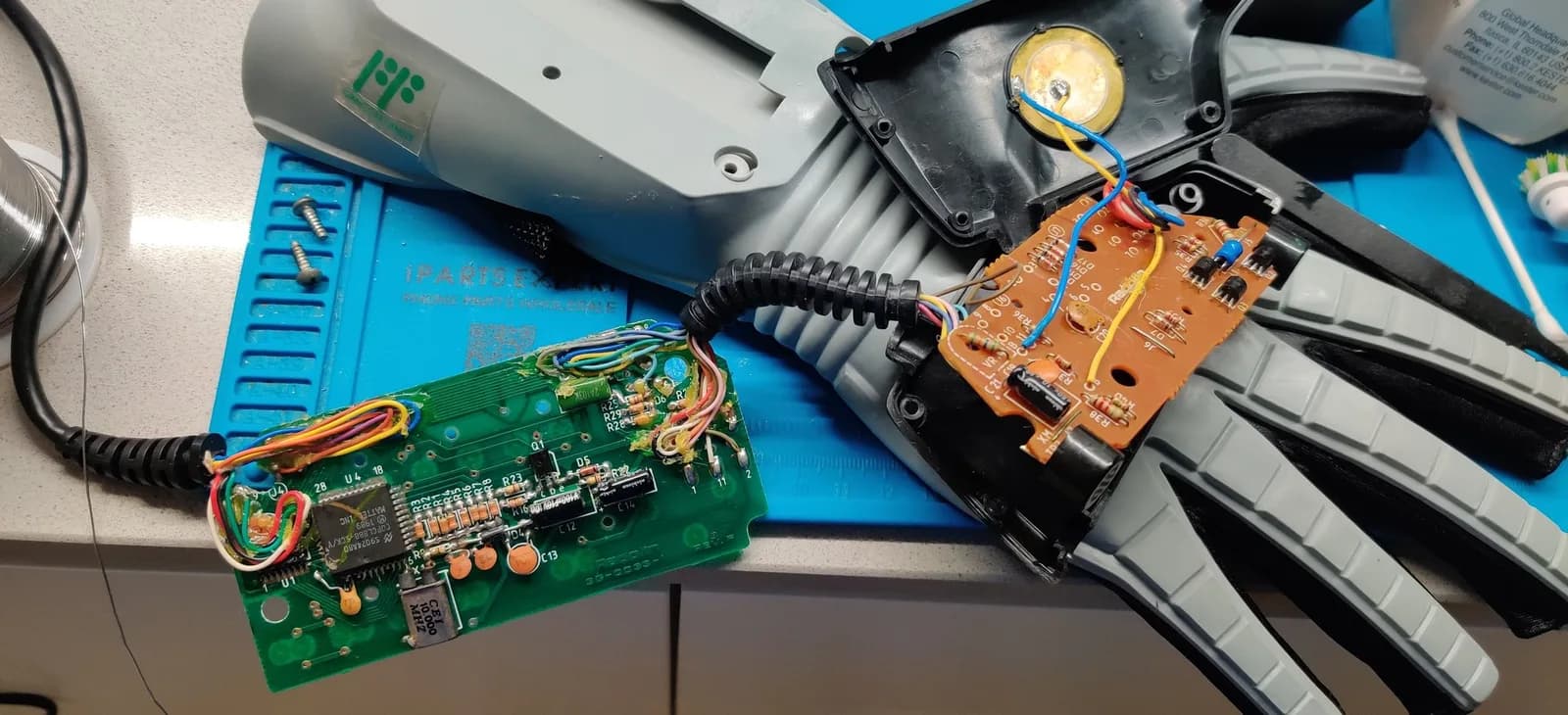 Nintendo Power Glove boards with the new capacitors now fitted: the green main PCB (square IC and a metal-can crystal at the edge) and the copper/orange daughterboard both recapped and joined by a colourful wire loom, alongside the grey glove internals, the black corrugated wrist cable and the round gold disc, on a blue work mat.