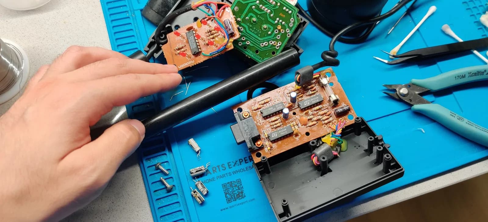 A hand holds a black plastic tube from the Power Glove assembly, the conduit that routes the wiring between the two square receiver boxes (the units that pick up the glove's input and hold the PCBs). Below sit the copper/orange board (two ST-marked ICs and a black connector) and the green board, with turquoise Xcelite 170M flush cutters, cotton swabs and small screws on the blue work mat.