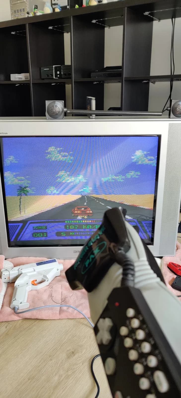 A powered-on Nintendo Power Glove held up in the foreground and tilted to the left, steering the car to the left in Rad Racer on the NES. The glove's square signal-receiver boxes sit on top of the Sony Trinitron KV-29FX30E CRT showing the game.