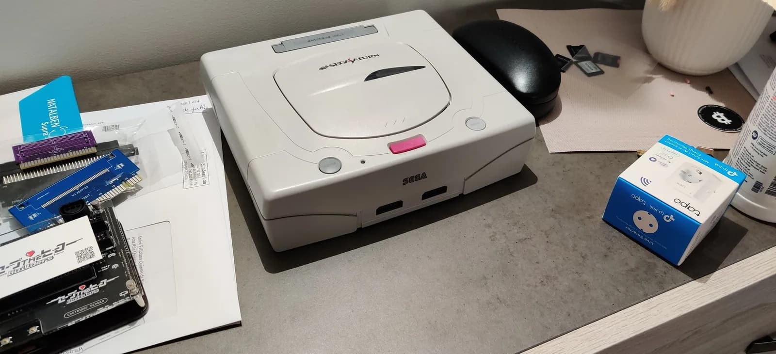 Retrobrited Sega Saturn model 2 on a desk, the plastic now almost back to its normal white, beside a prescription-glasses case, with documentation and spare boards nearby.