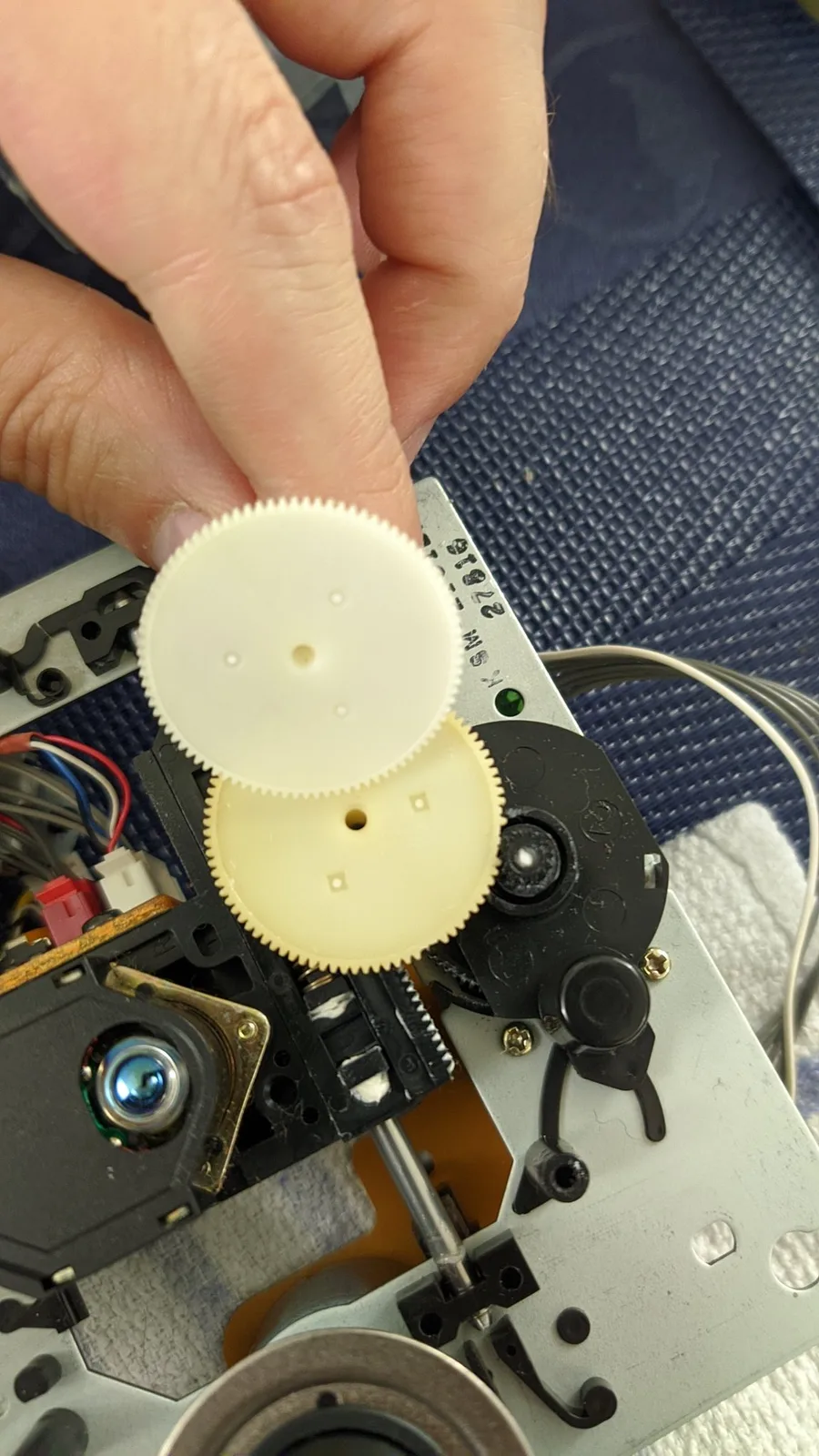 Hand holding a new replacement gear (all teeth intact) for the NEC PC Engine Super CD-ROM2 beside the old yellowed gear, which is missing several teeth, during the refurbishment.
