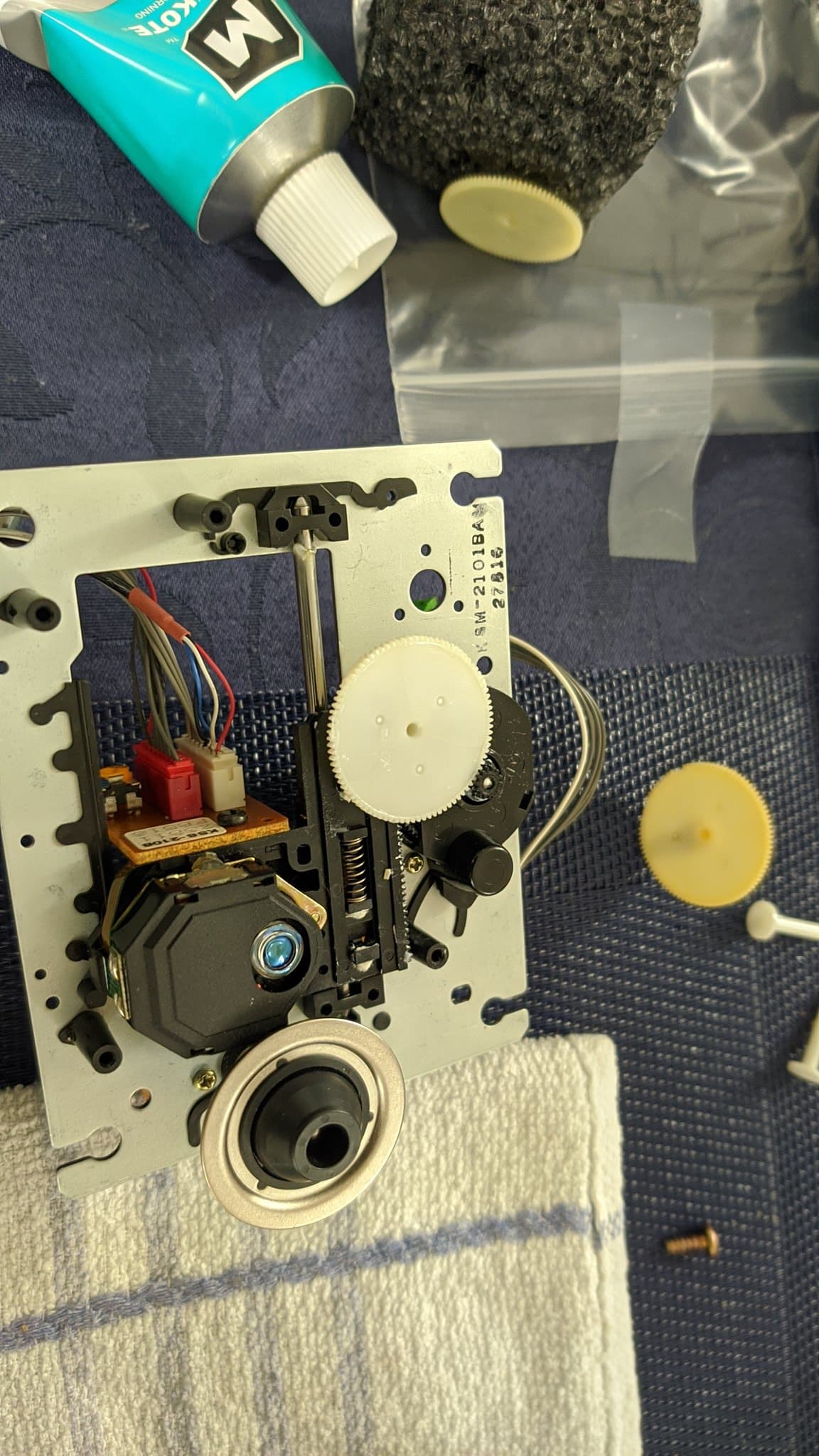 The NEC PC Engine Super CD-ROM2 laser assembly with the new gear installed, the old gear on the table beside it and a tube of Molykote nearby, used to lubricate the moving parts of the laser sled.