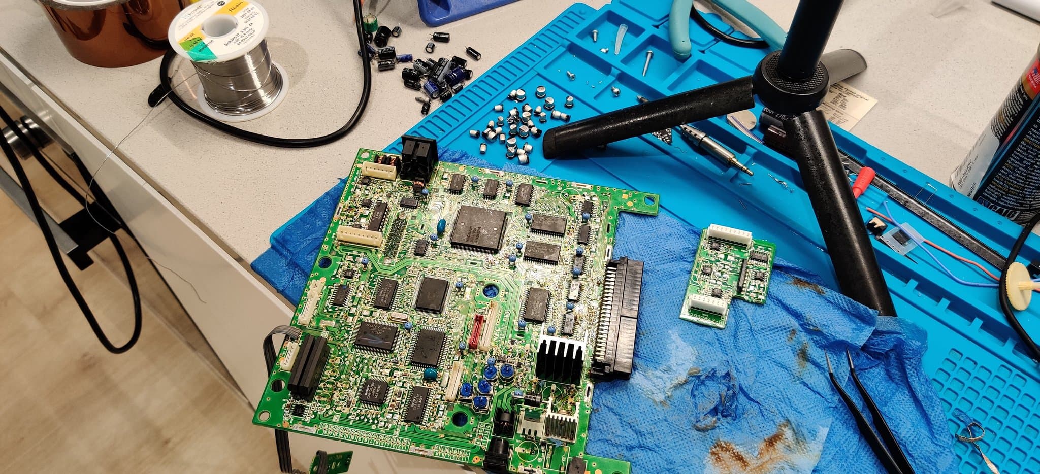 NEC PC Engine Super CD-ROM2 with both boards fully depopulated, the mainboard and the daughterboard, their old capacitors removed and laid out on the blue mat alongside desoldering braid.