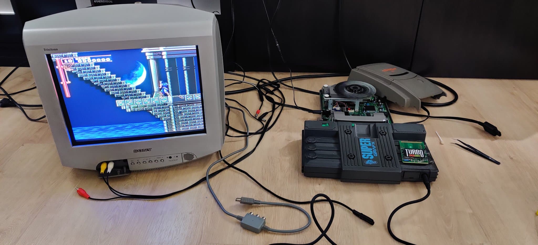 NEC PC Engine Super CD-ROM2 connected to a CRT displaying Castlevania: Rondo of Blood gameplay, the recapped motherboard visible, with a pair of tweezers and the unit's shell on the floor beside it during the refurbishment.