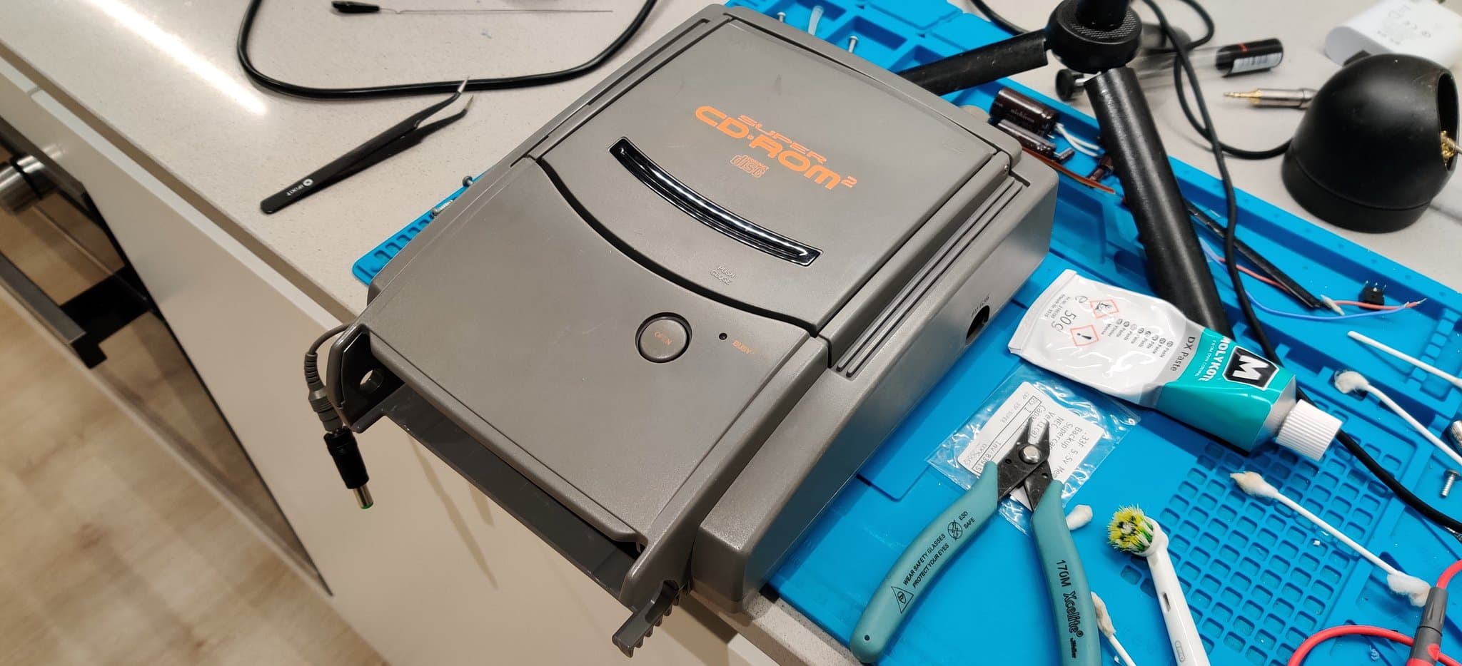 NEC PC Engine Super CD-ROM2 unit on the work mat after retrobrighting, alongside a tube of Molykote, a pair of tweezers, a pair of pliers and a toothbrush head used for cleaning.