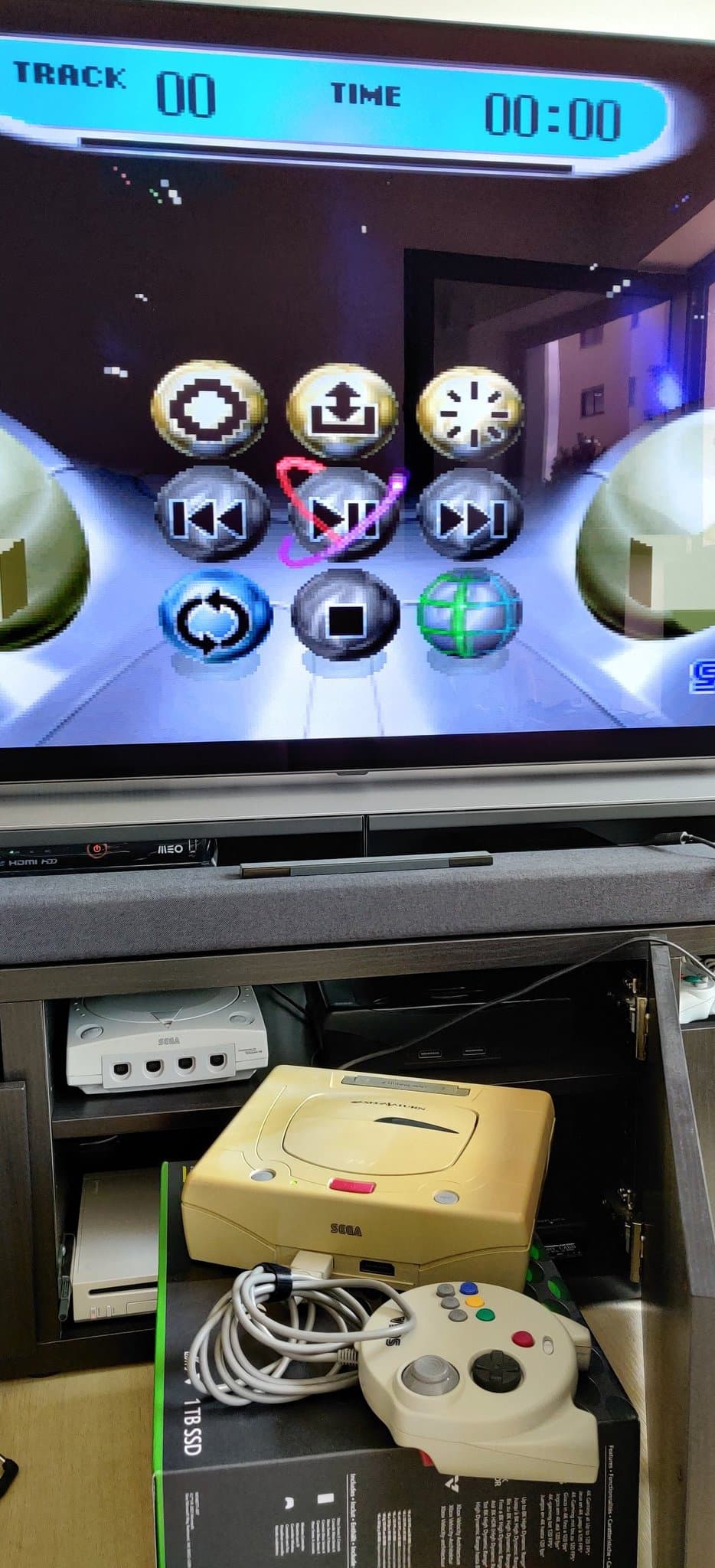 Sega Saturn yellow Model 2 console displaying media player menu on TV, with controller and cables visible below in storage drawer.