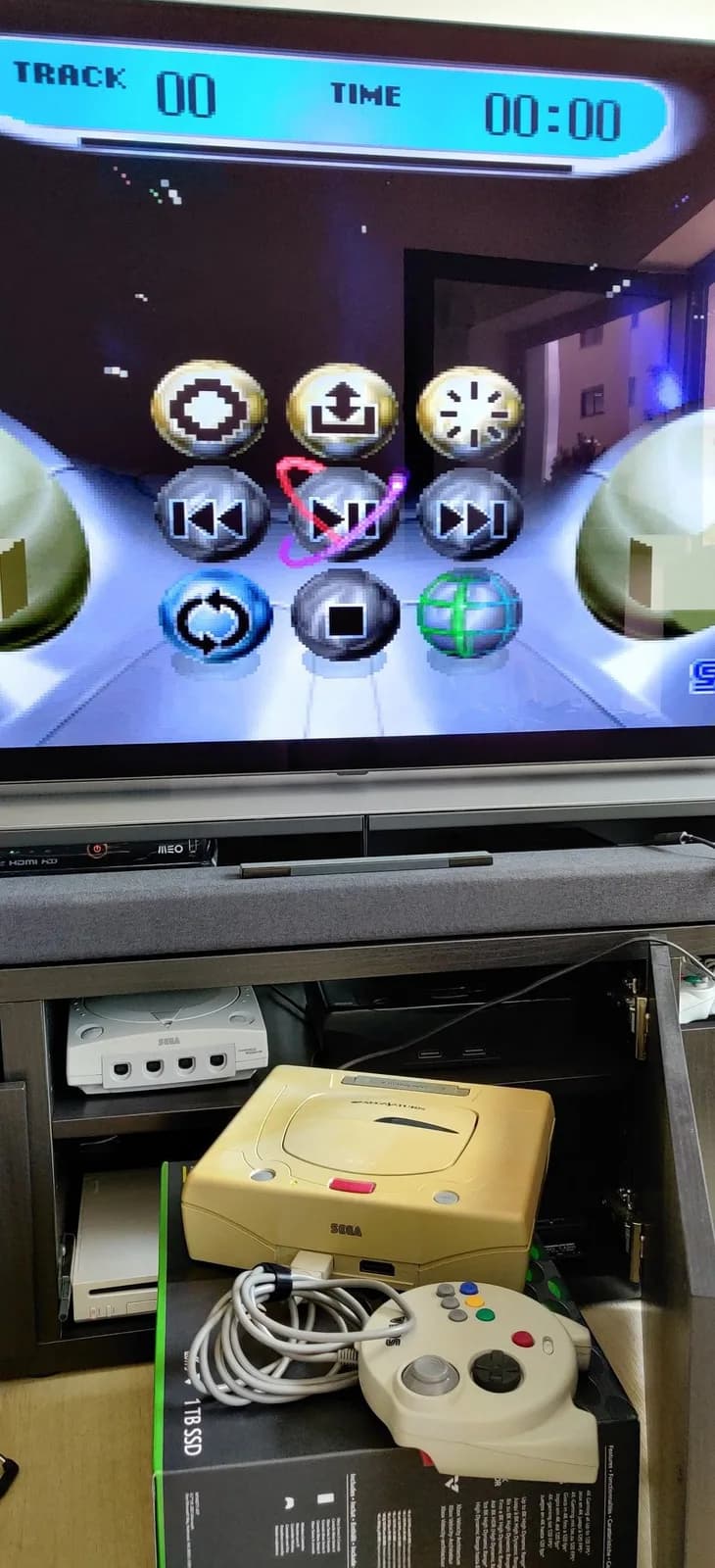 Sega Saturn yellow Model 2 console displaying media player menu on TV, with controller and cables visible below in storage drawer.