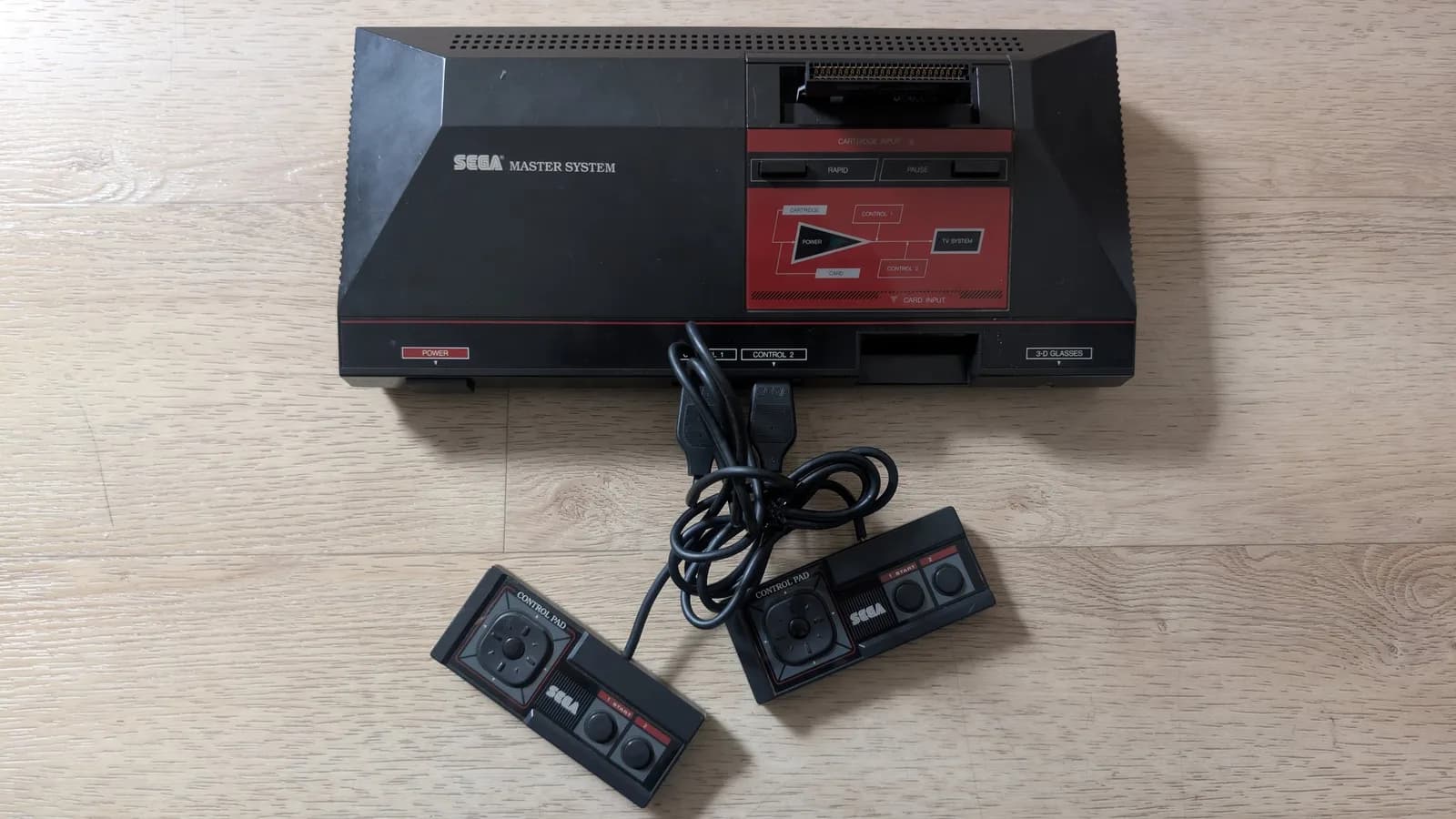 Sega Master System MK-2000 with two controllers connected via cable, displayed on wooden surface during refurbishment.