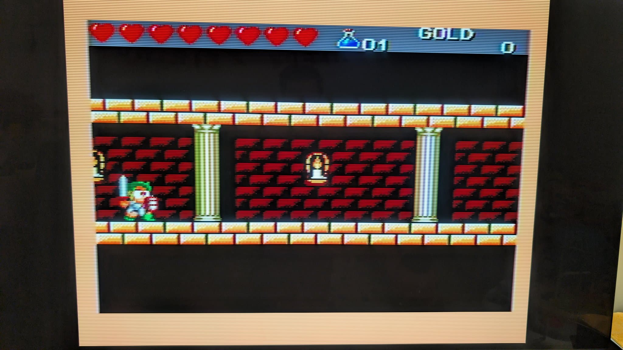 Master System displaying Monster Boy III gameplay: a small green-clad hero holding a sword and shield stands on red-brick flooring between stone columns, with a full row of red hearts and a GOLD counter along the top of the screen.